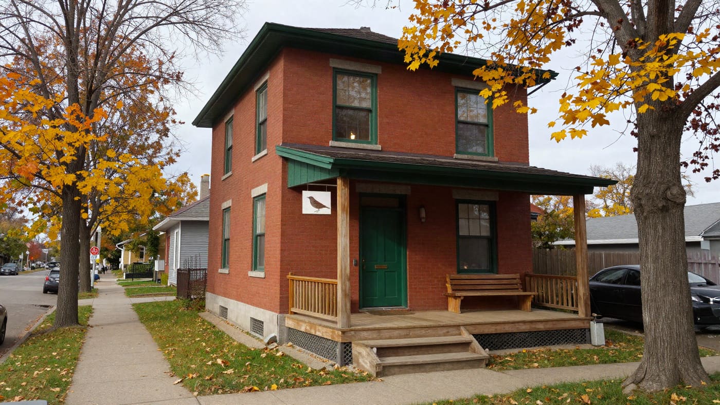 The red-brick house with a green door and a painted wren sign over the porch, autumn trees